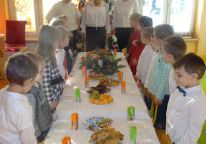 Pani dyrektor Maria Królikowska, pani Agnieszka Piekarska, pani Arleta Kalinowska oraz dzieci stoją wokół nakrytego białym obrusem i poczęstunkiem stołu.